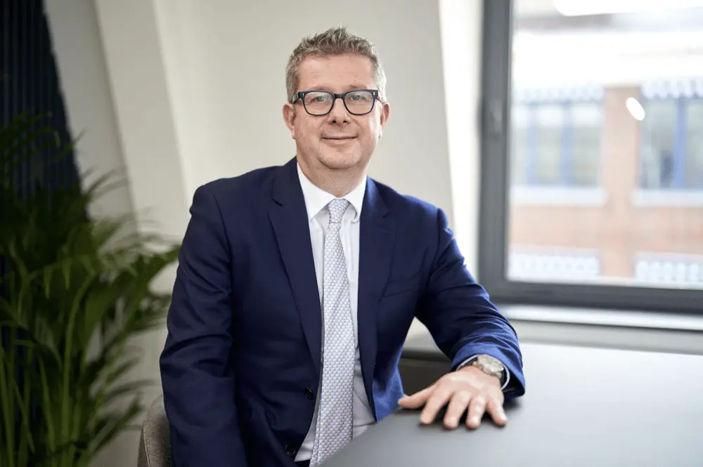 Un homme en costume bleu assis dans un bureau moderne de Shaftesbury Capital, souriant légèrement.