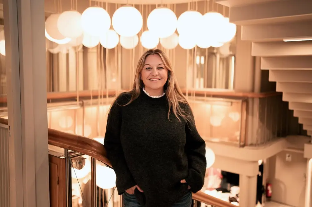 Une femme souriante pose sur un balcon intérieur éclairé par des lampes rondes, représentant une ambiance chaleureuse chez Shein