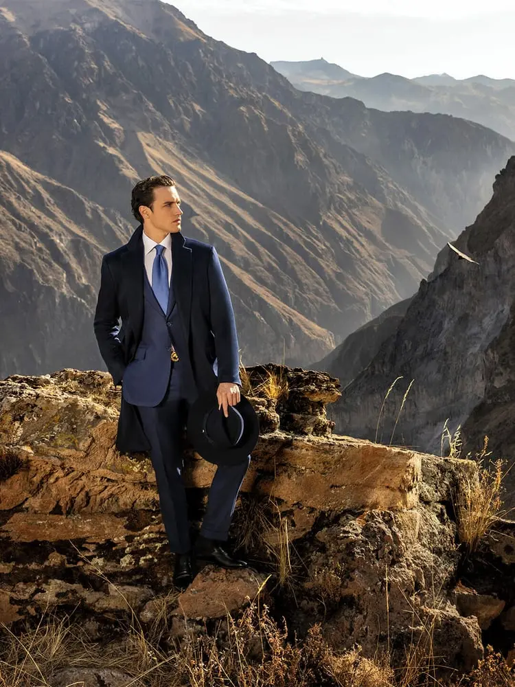 Homme élégant en costume Stefano Ricci tenant un chapeau, se tenant sur un rocher avec vue sur montagne majestueuse