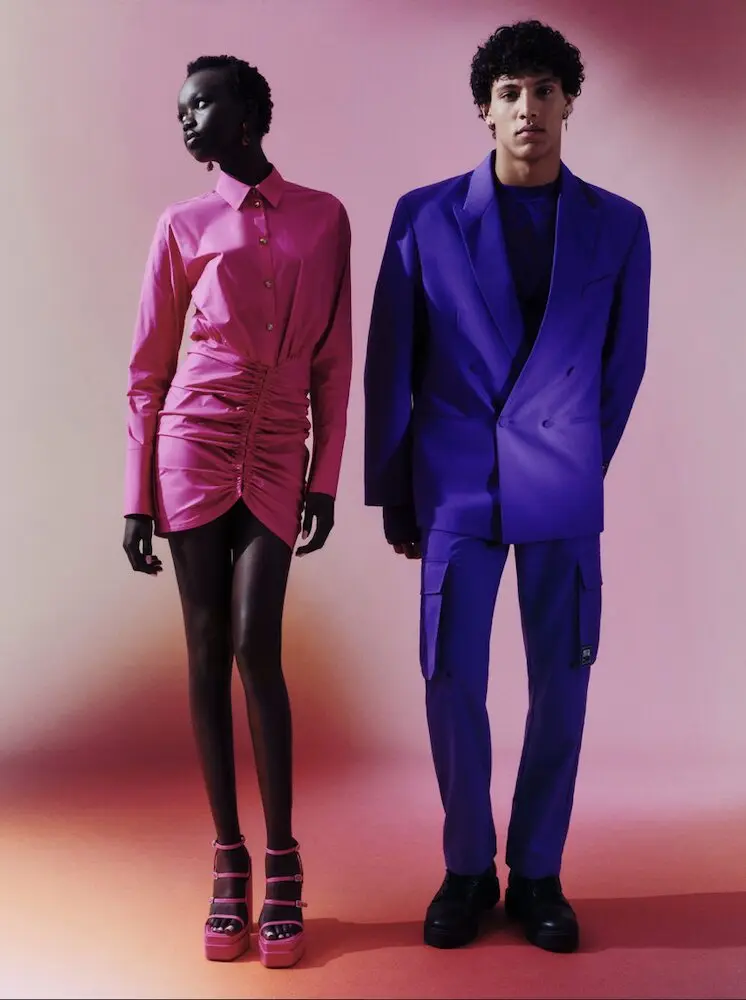 Deux mannequins posent en tenues colorées, l'un en robe rose et l'autre en costume violet, fond dégradé.