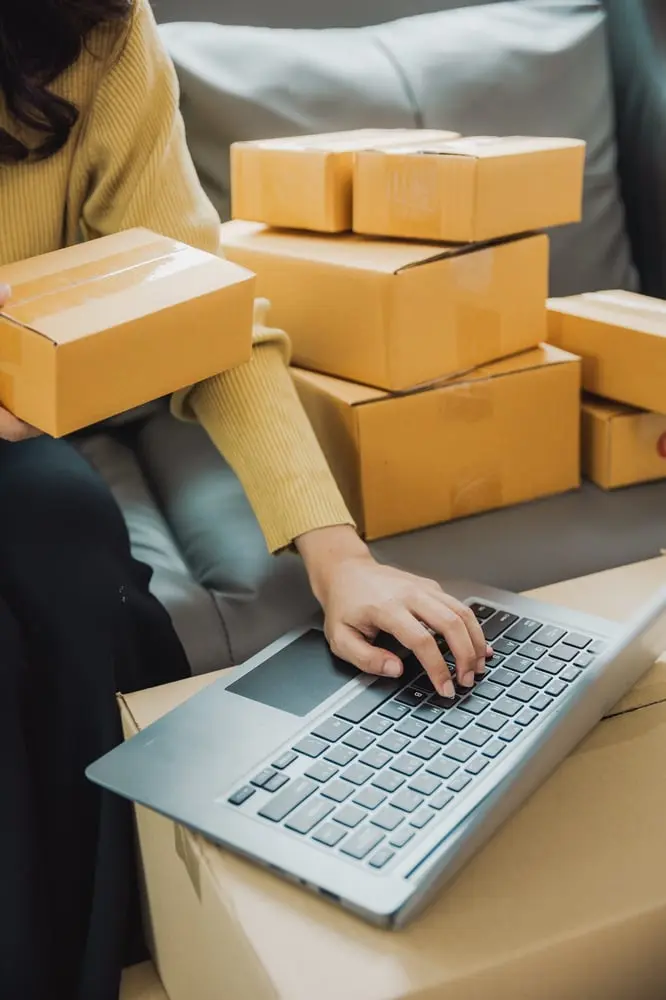 Personne en pull jaune utilisant un ordinateur portable sur une pile de colis, illustrant l'impact de la taxe de 3 €