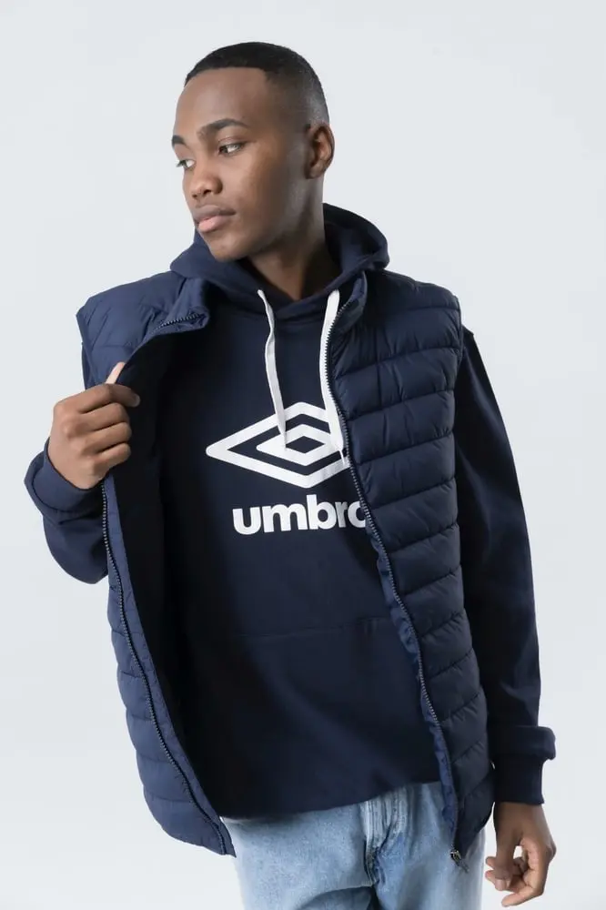 Un homme porte un sweat bleu avec le logo Umbro et une veste sans manches assortie.
