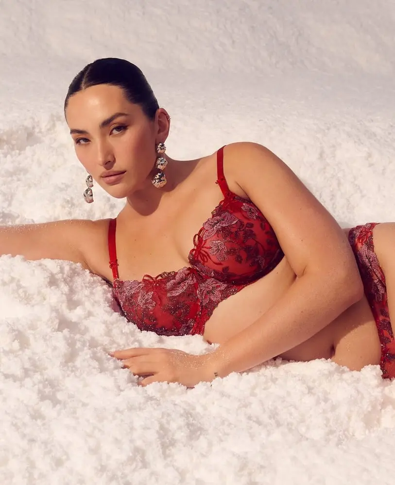 Modèle en lingerie rouge Victoria’s Secret posant allongée dans la neige, portant de longs bijoux d’oreilles.
