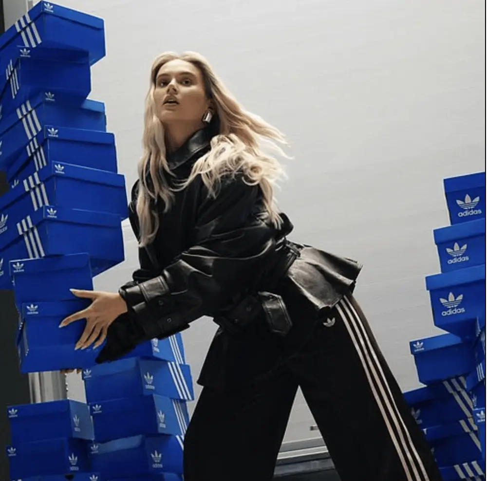 Femme tenant des boîtes bleues Adidas empilées, portant un manteau en cuir, cheveux blonds flottants.