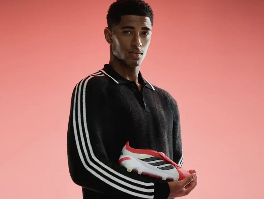 Un homme en pull Adidas noir tient une chaussure de football Predator sur fond rose