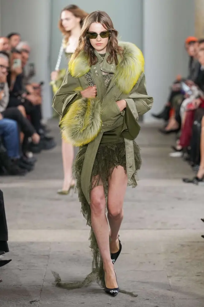 Mannequin défilant en manteau vert avec fourrure jaune vif Blumarine sur un podium rempli de spectateurs.