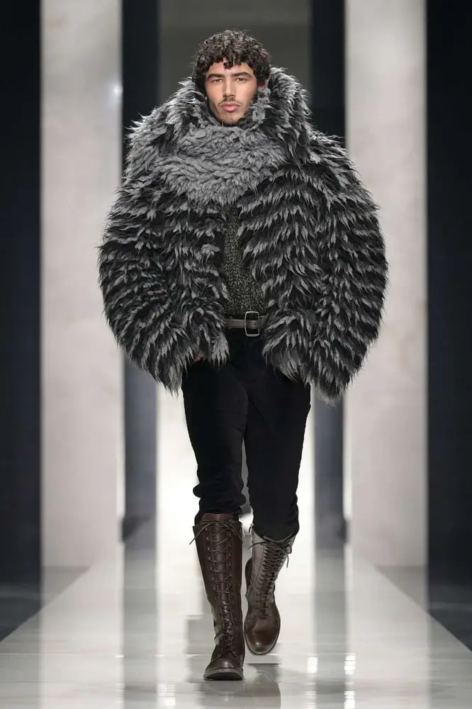 Modèle homme défilant à Milan pour Dolce & Gabbana en 2026, portant un manteau en fausse fourrure grise et des bottes hautes.