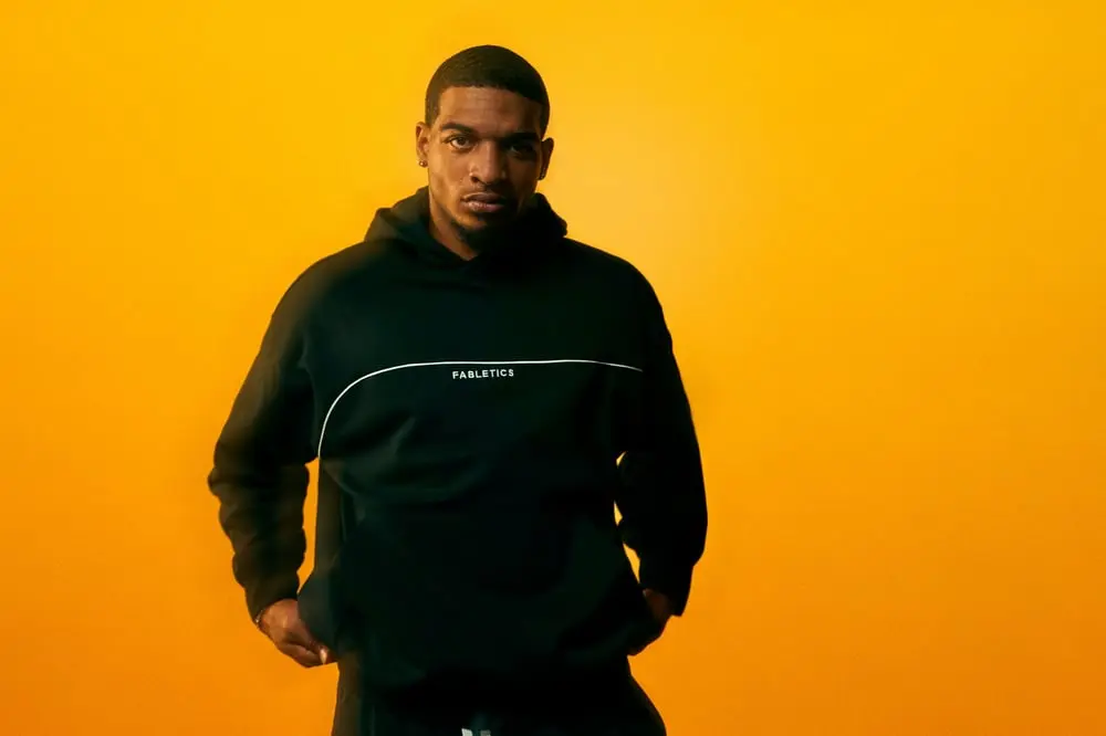 Un homme en sweat noir Fabletics pose devant un fond orange, représentant la nouvelle collection homme