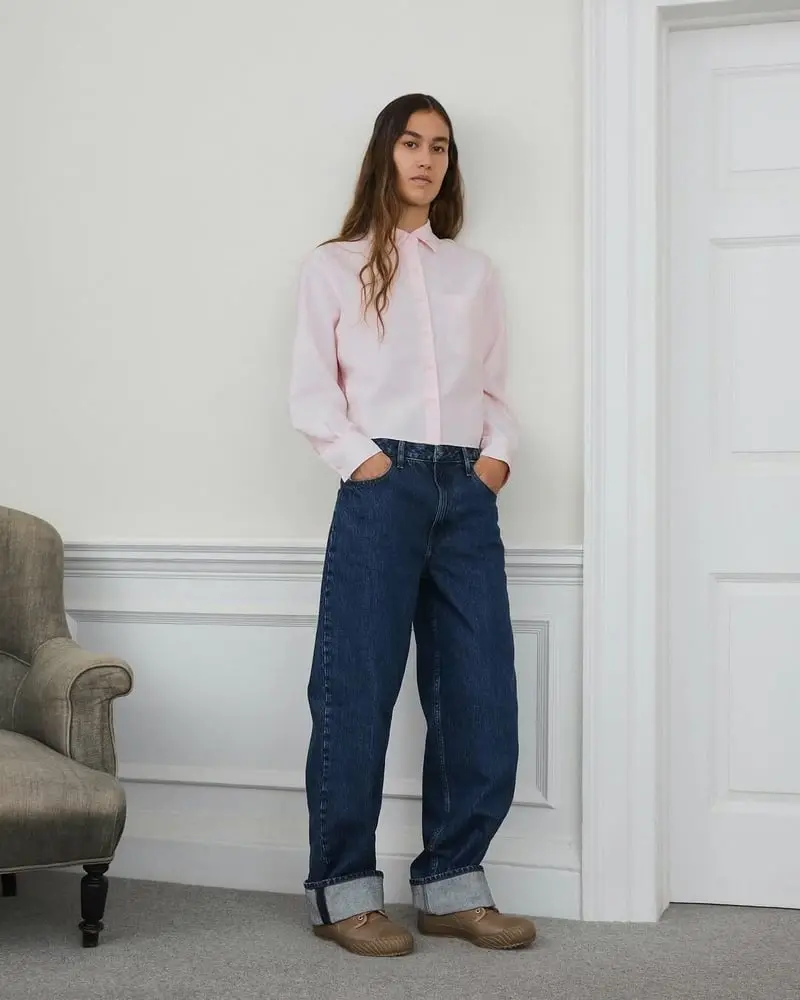 Personne portant une chemise rose et un jean ample, debout dans une pièce au décor épuré, évoquant le style Uniqlo