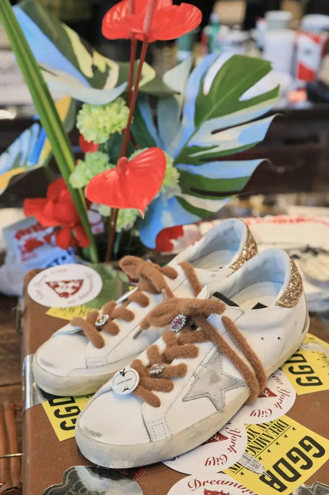 Des baskets Golden Goose blanches et dorées avec des lacets marron, décorées de badges, posées sur une boîte avec des autocollants.