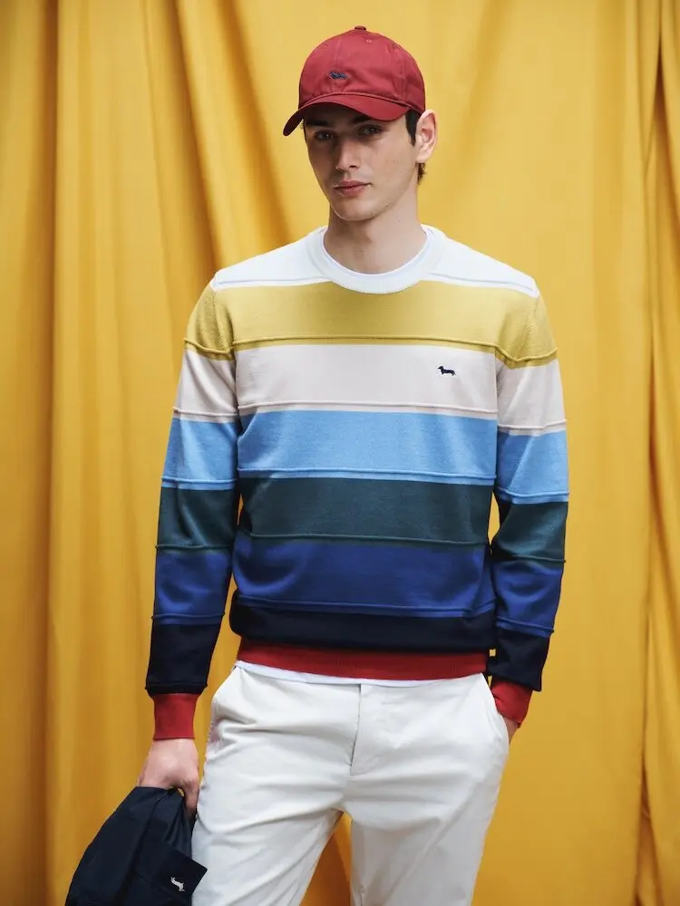 Un homme en pull rayé coloré et casquette rouge pose devant un fond jaune pour Harmont & Blaine