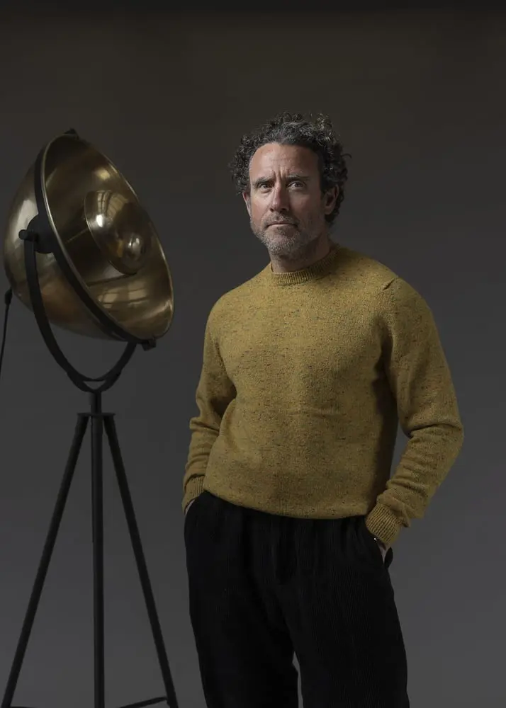 Un homme en pull ocre se tient devant un grand projecteur, représentant le style innovant de la marque Inis Meáin