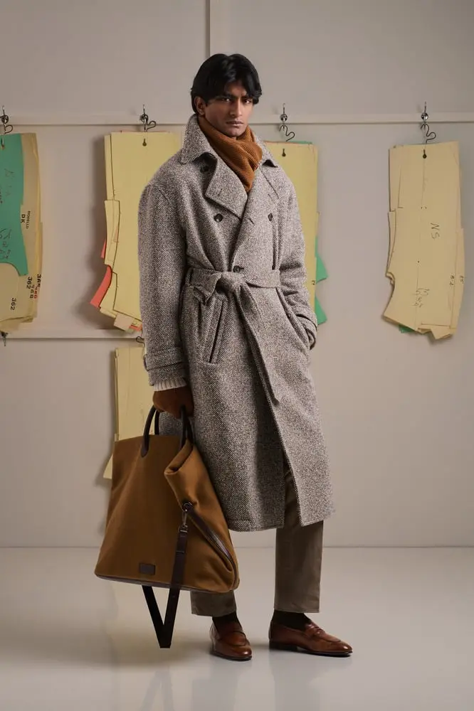 Un homme en manteau gris Kiton pose devant des patrons de couture, tenant un grand sac marron