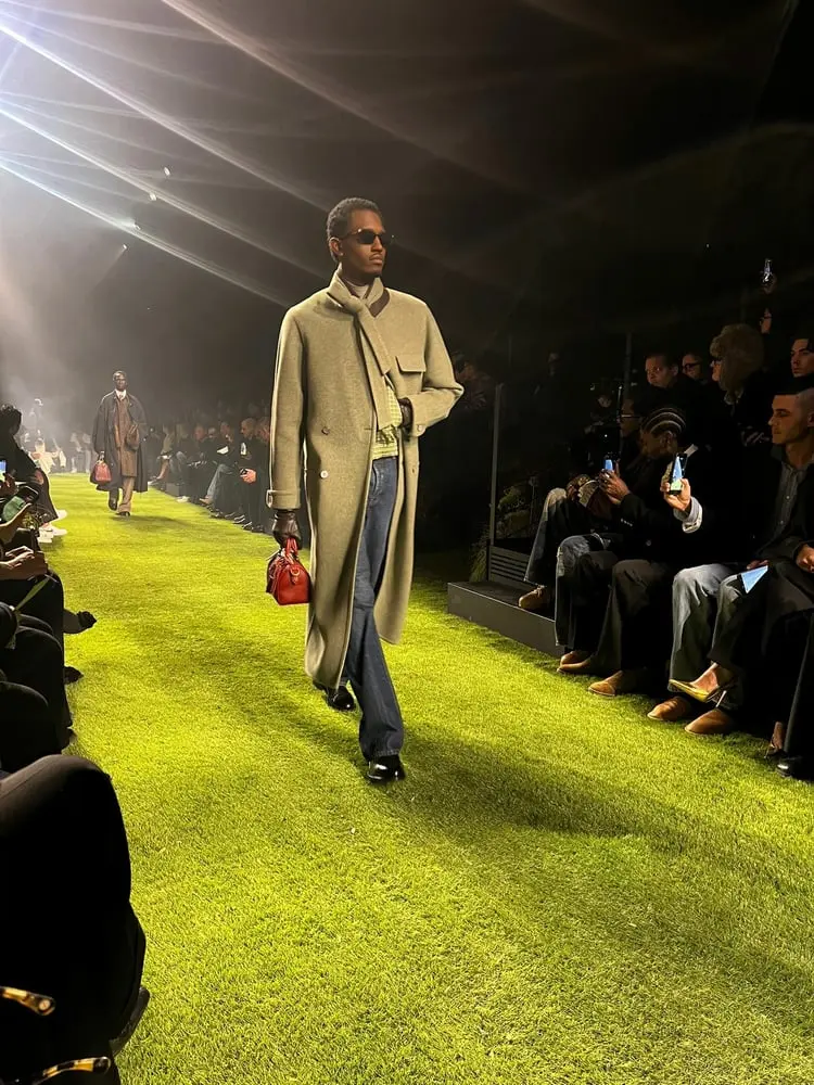 Modèle défilant sur un podium vert lors de la Paris Fashion Week Homme de Louis Vuitton, portant un manteau élégant.