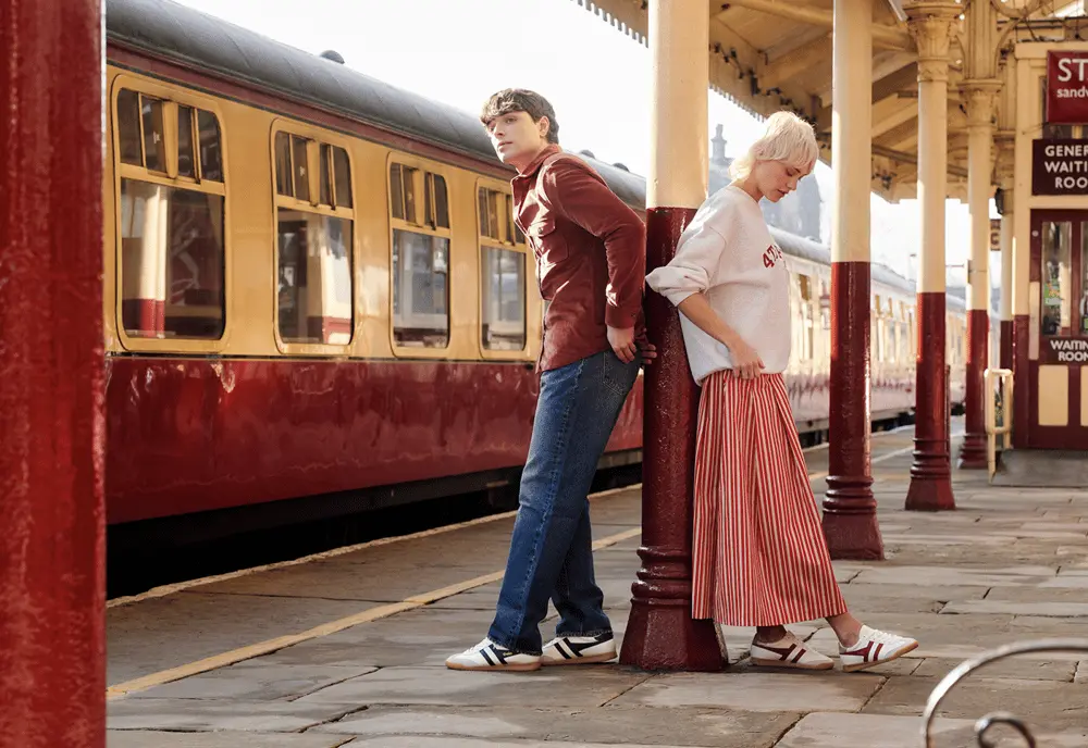 Des personnes portant des chaussures Gola posent sur un quai de gare vintage, appuyées contre un pilier.
