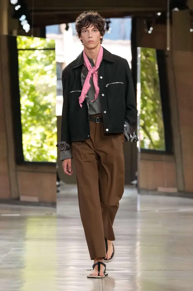 Un mannequin marche sur le podium de la Fashion Week à Paris, vêtu d'un ensemble chic et décontracté avec foulard rose.