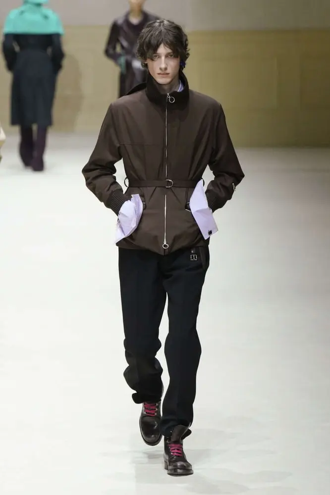 Un mannequin défile en tenue Harrods avec une veste marron et pantalon noir, illustrant la tendance luxe réfléchi.
