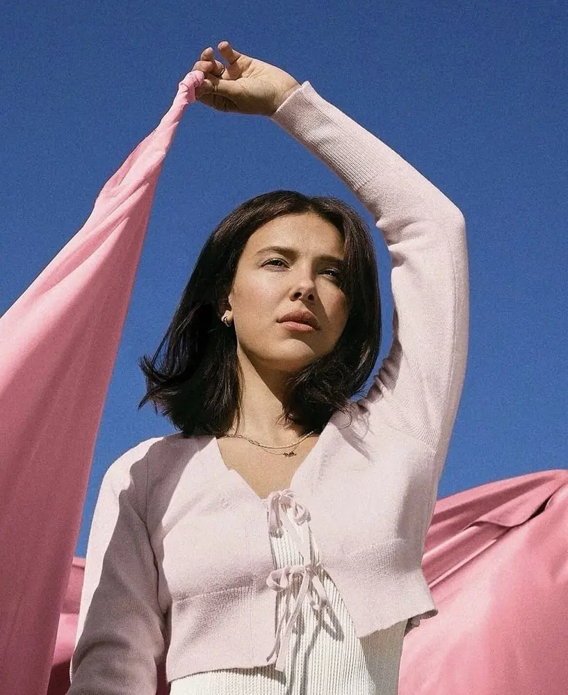Une jeune femme en haut rose tient un tissu rose devant un ciel bleu, illustrant la marque Mills chez Walmart