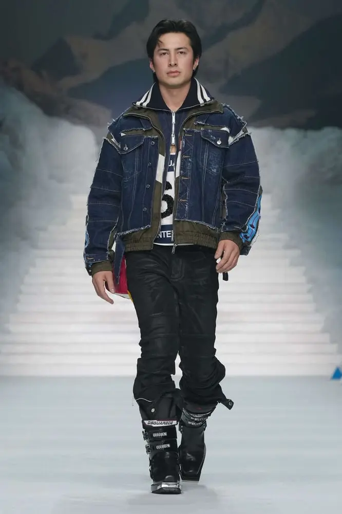 Un homme défile à Milan en tenue Dsquared2 avec veste en jean et bottes noires devant un décor montagneux.