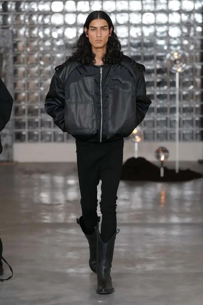 Un mannequin défile à Milan portant une veste en cuir noir Orefice et des bottes dans un cadre industriel.