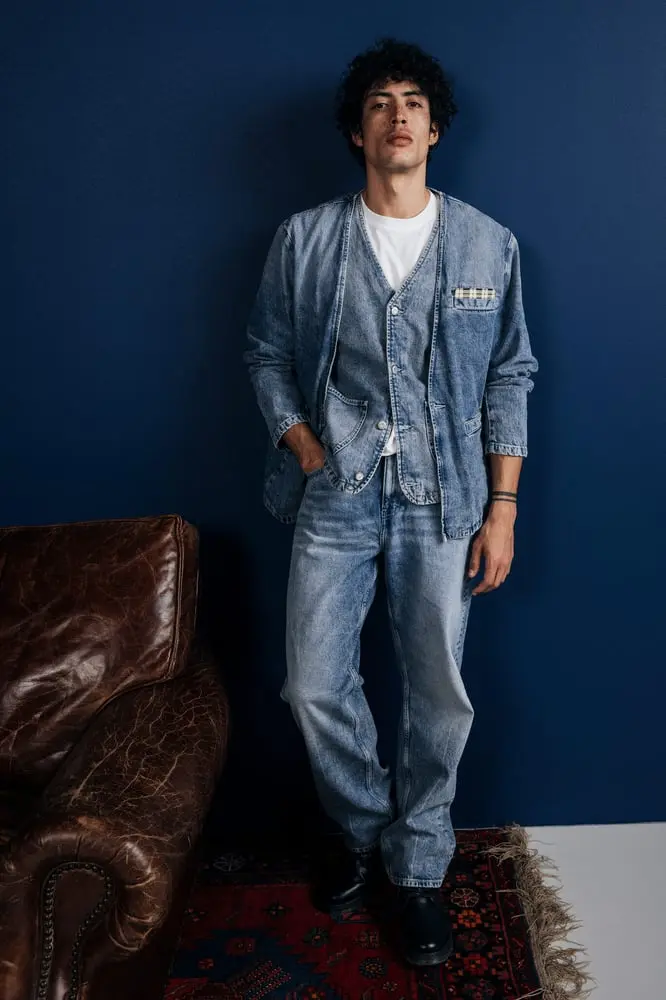 Un homme en jean et veste en denim Mustang pose devant un mur bleu avec un canapé en cuir et un tapis coloré