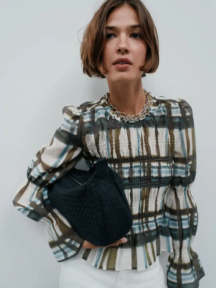 Femme portant une blouse à carreaux et un sac en cuir tressé de Zalando, posant devant un mur clair