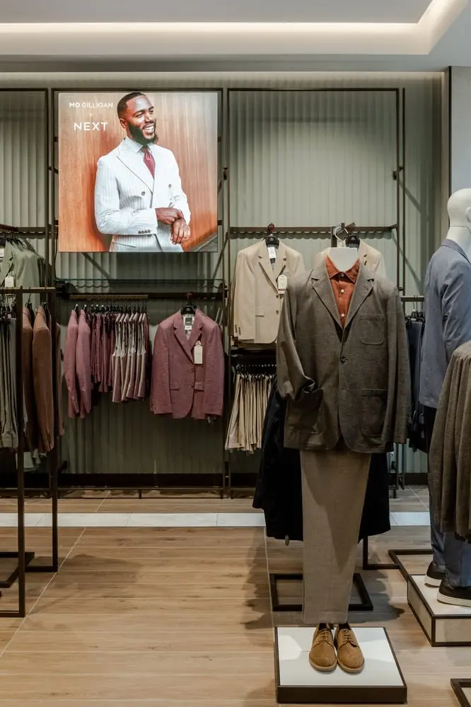 Magasin Next avec costumes sur des mannequins et affiche d'un homme souriant en costume sur le mur.