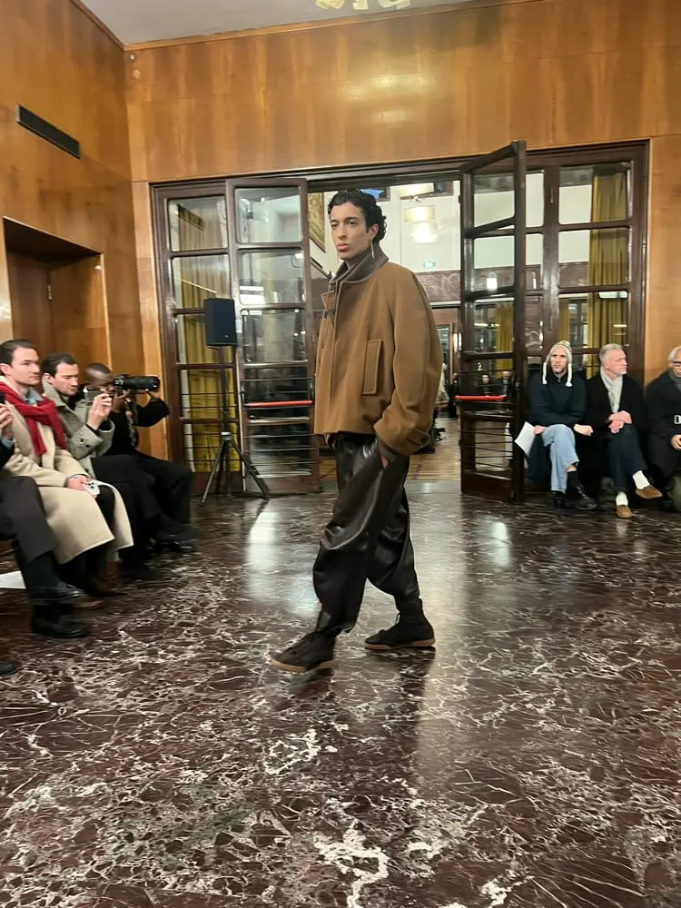 Défilé de mode masculine Pitti Uomo 2025 avec un mannequin en veste marron et pantalon large brun dans une salle aux murs en bois