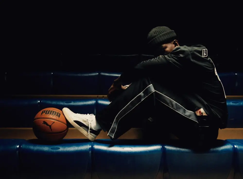 Un homme assis dans des gradins sombres avec un ballon de basketball Puma, portant des vêtements de sport élégants