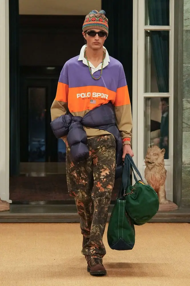 Modèle portant une tenue Ralph Lauren mixant style sportif et motifs camouflages, avec un pull violet et orange Polo Sport