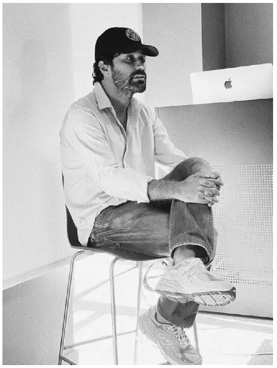 Un homme assis sur un tabouret portant une casquette et des baskets dans un bureau avec un ordinateur portable Sporty & Rich