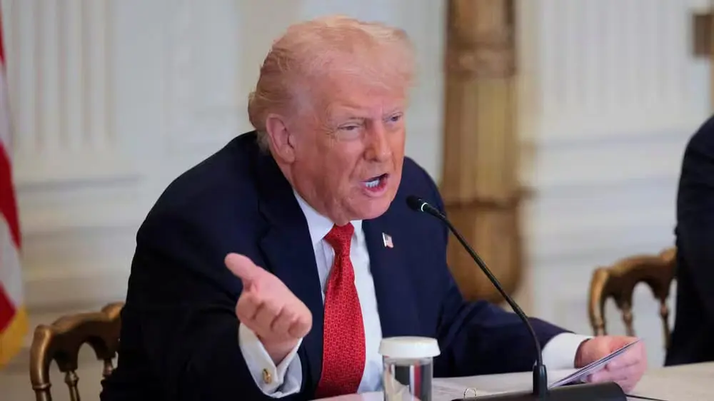 Je ne sais pas qui est la personne sur l'image. Un homme parle dans un micro à une table, gesticulant avec la main dans une salle ornée.