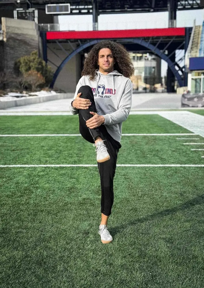 Un homme en tenue décontractée sur un terrain de sport effectue un étirement, illustrant la collaboration Vivobarefoot