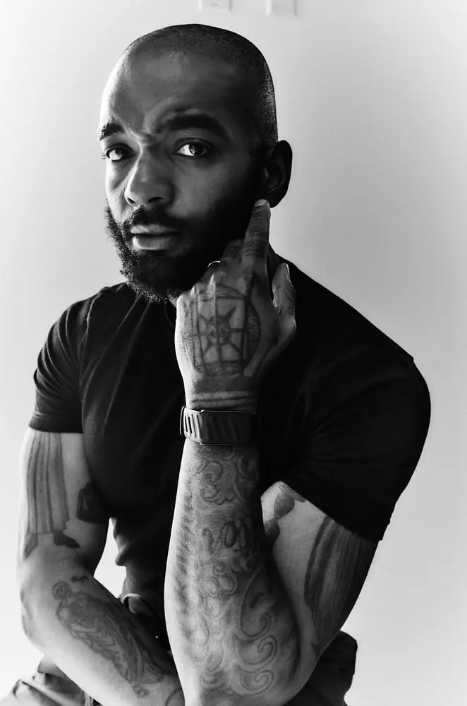 Homme tatoué en t-shirt noir portant une montre, posant pensivement pour annoncer le partenariat Whoop